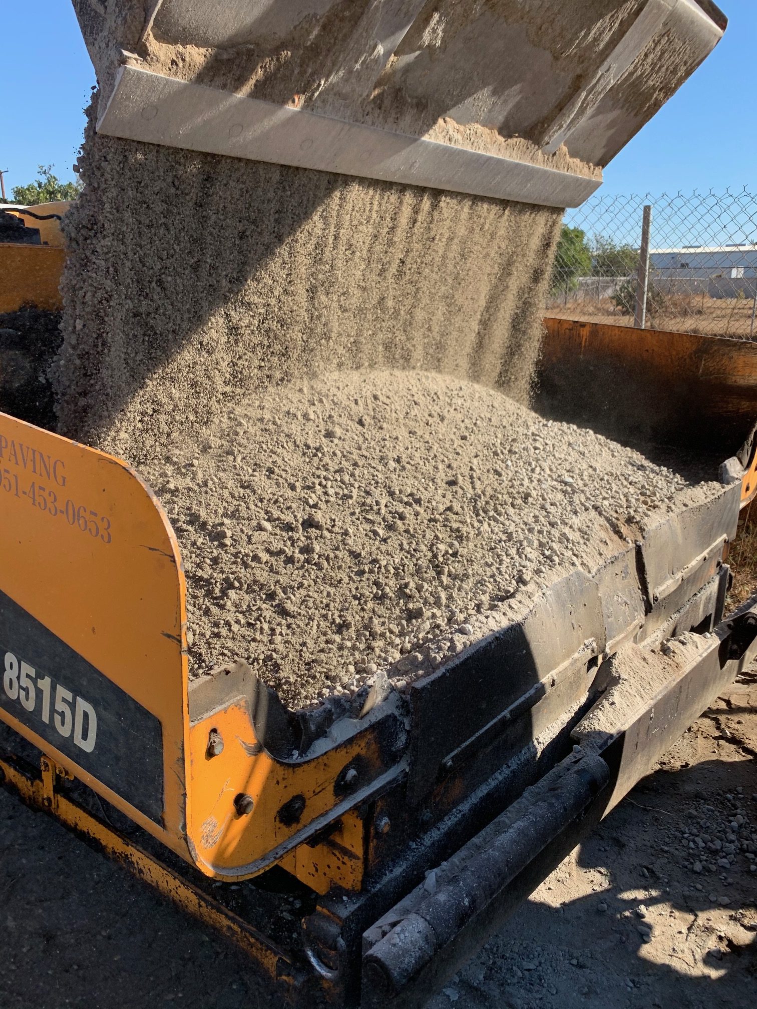 A machine pours gravel into a yellow-black paving vehicle